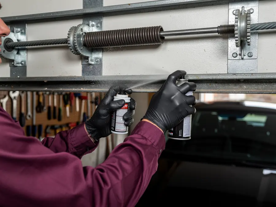 A technician in black gloves sprays lubricant on a garage door torsion spring.